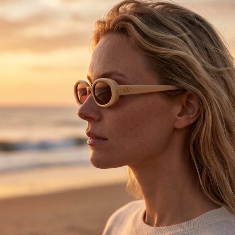 Femme à la plage portant les lunettes de soleil crème Le Specs Outta Love face au coucher du soleil.